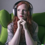 A red-haired, green-eyes young adult woman wearing big headphones and sitting in a green wingback armchair