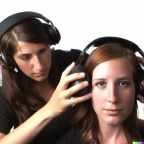 A photograph of an attractive young woman wearing large black vintage headphones, putting large black vintage headphones over the ears of another attr