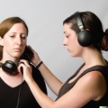 A photograph of an attractive young woman putting large black vintage headphones onto the ears of another attractive young woman, high resolution, wid
